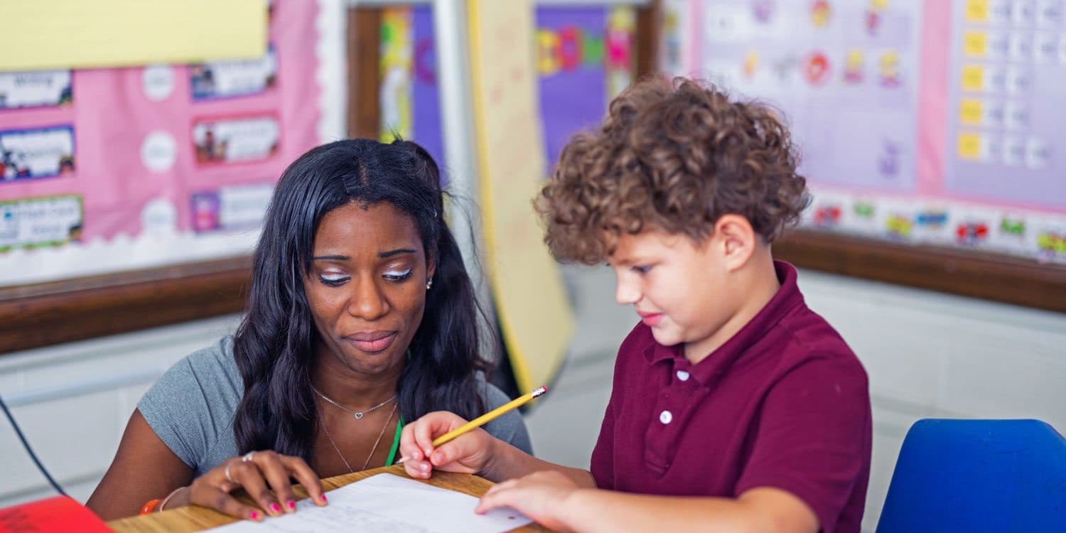 An adult working one-on-one with a student as the student writes at a classroom desk.